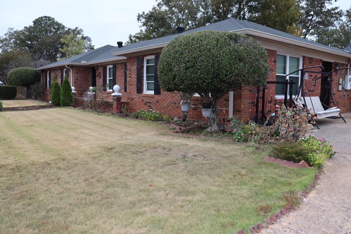 A single-story brick building is surrounded by well-maintained lawns and shrubs. The property features potted plants and a swing set, providing an inviting outdoor space for relaxation. Windows are adorned with white shutters, contributing to the overall charm of the home.
