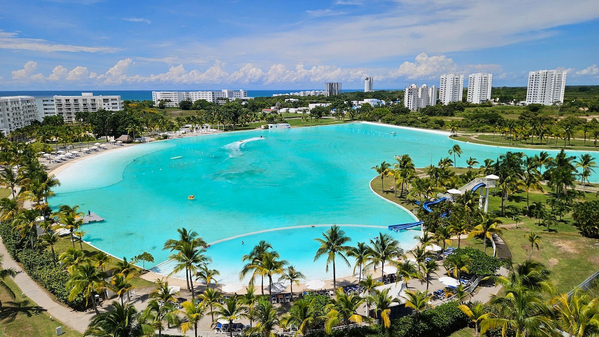 A large saltwater pool is prominently displayed, bordered by lush greenery and palm trees. Sun loungers are arranged along the sandy shore. In the background, several tall buildings overlook the shimmering water, and the vast Pacific Ocean stretches into the horizon under a clear blue sky.