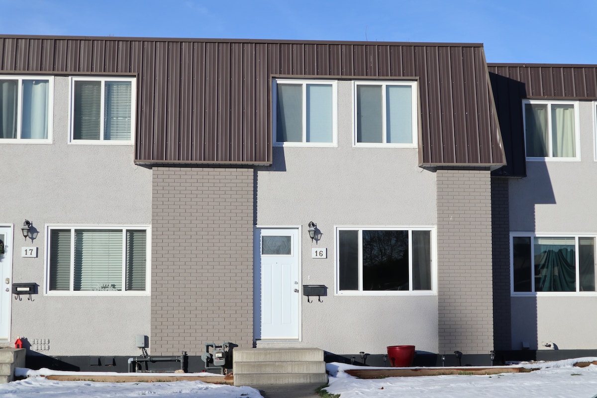 The exterior of a two-story townhome is shown, featuring a modern design with large windows. A white front door is centered, flanked by light fixtures. The surrounding area has patches of snow, and there are steps leading to the entrance. The building is part of a row of similar units.