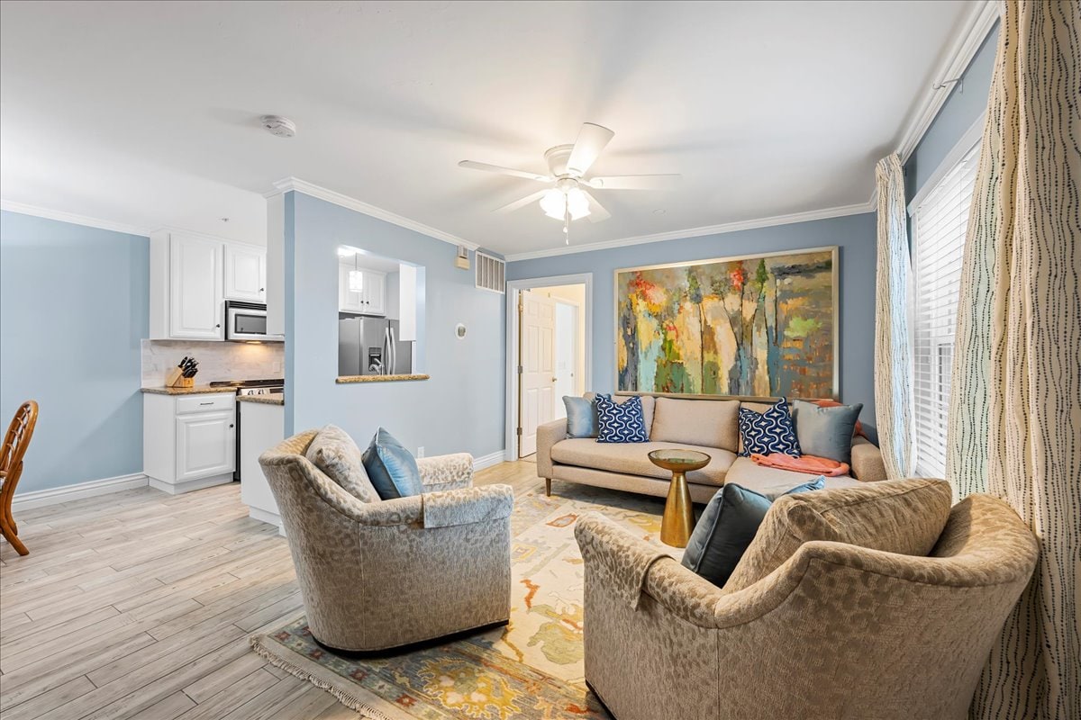 A spacious living area features a light blue color scheme, with a large abstract painting on the wall. Comfortable seating is provided by two upholstered armchairs and a sofa. A wooden side table rests between the chairs, while natural light streams in through the window.
