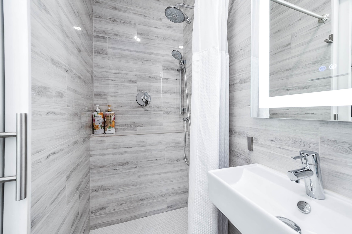 A modern bathroom is presented with a sleek shower area featuring dual shower heads. Light gray wood-look tiles adorn the walls, complementing a white vanity with a polished faucet. A large mirror enhances the space, while toiletries are neatly organized on a wall shelf.