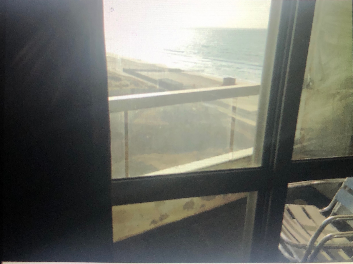 A view from a balcony showcases a serene coastal landscape, with the ocean waves gently lapping at the shore in the distance. The glass paneling allows for unobstructed views of the sandy beach and tranquil waters, with sunlight reflecting on the surface.
