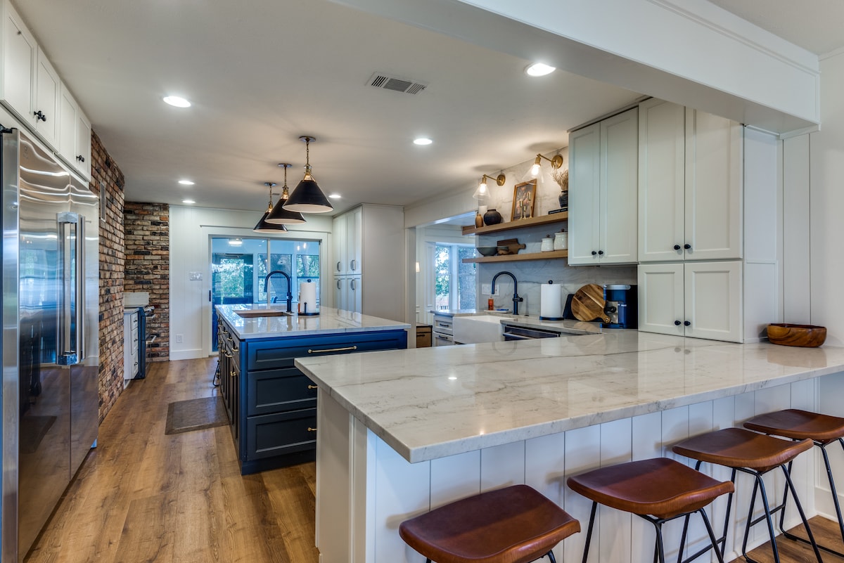 A spacious kitchen features a large island with seating, complemented by modern bar stools. A mixture of white cabinetry and blue accents enhances the design, while stainless steel appliances and a rustic brick wall add character. Natural light pours in through large windows, illuminating the area.