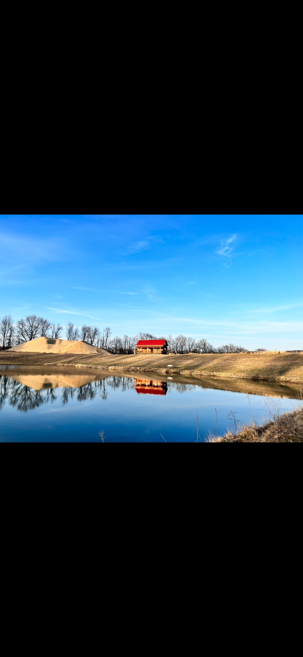 A rustic cabin stands beside a tranquil pond, its red exterior reflected in the calm water. The surrounding landscape features gentle hills and a clear blue sky, contributing to a serene rural ambiance.