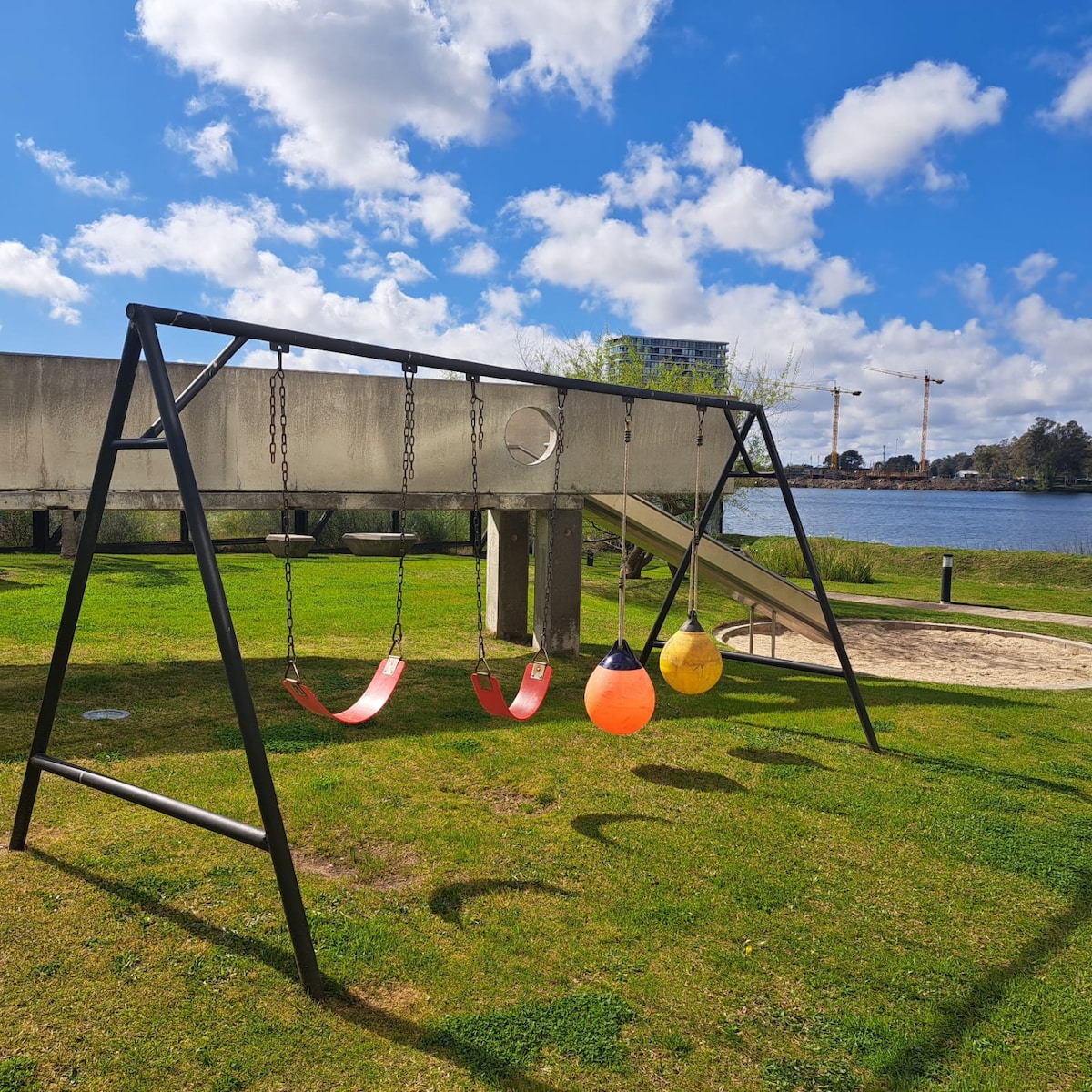 A playground area showcases a swing set with several colorful swings in red and orange, set against a backdrop of green grass. A slide is visible in the background, with the shimmering lake and blue sky enhancing the scene.