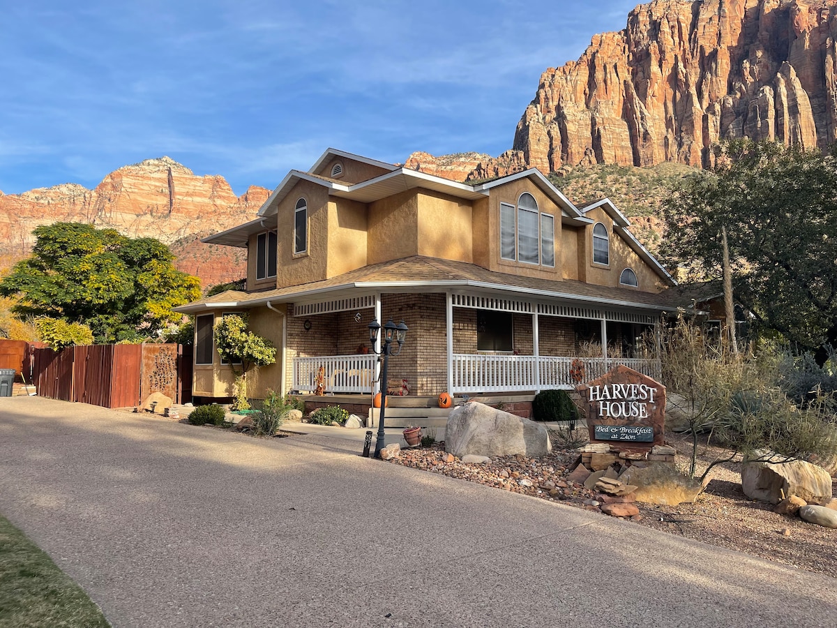 A two-story house is set against a dramatic mountain backdrop, featuring a welcoming front porch and decorative elements like lanterns. The warm exterior color complements the natural surroundings, while a landscaped area with various plants enhances the curb appeal.