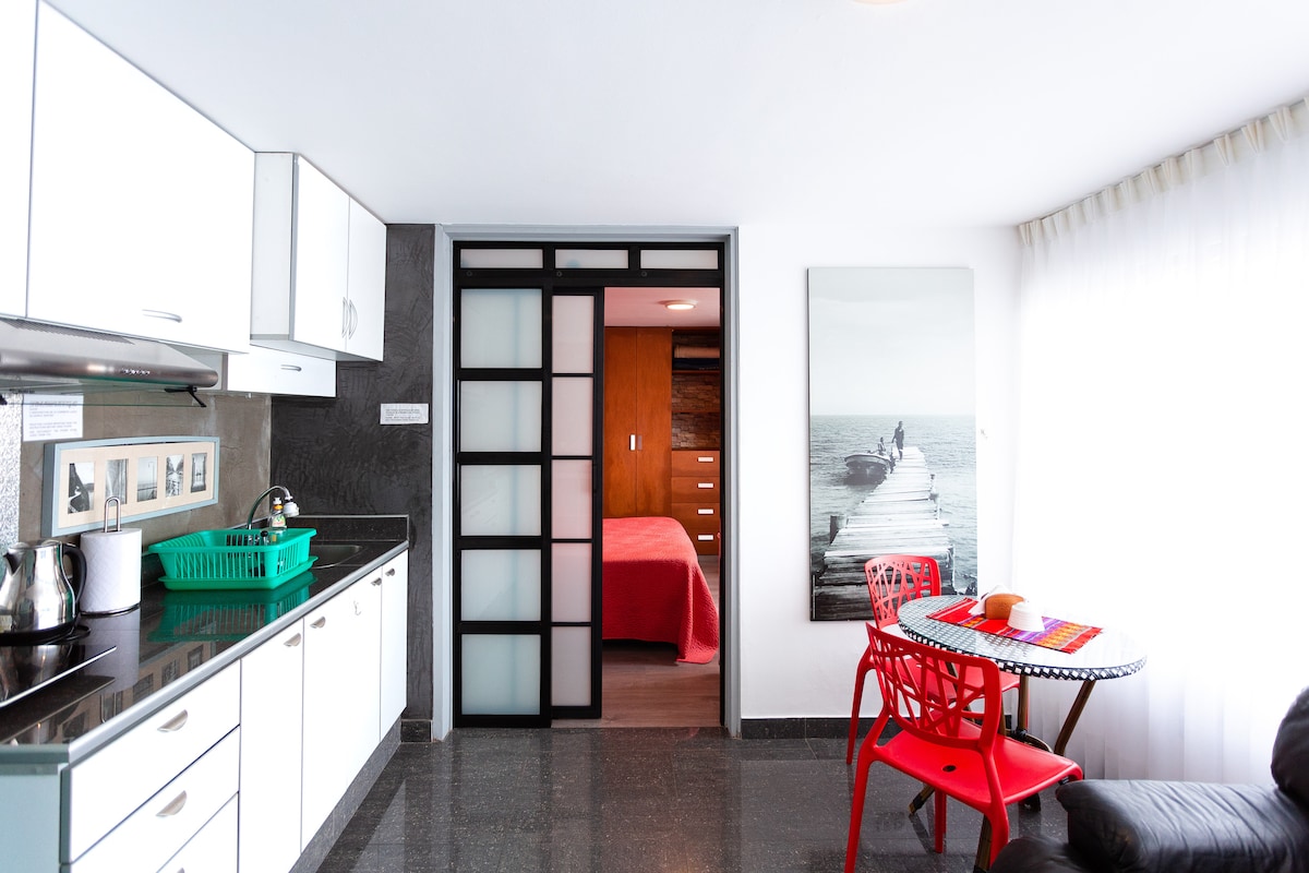 The modern kitchen features sleek white cabinetry and dark countertops, complemented by stainless steel appliances. A round dining table with red chairs is positioned nearby. A sliding glass door leads to a cozy bedroom area, while natural light filters through sheer curtains.