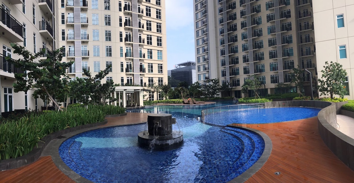 A large outdoor swimming pool occupies the center of the scene, featuring a curved shape with blue tiles. Surrounded by well-maintained greenery and two adjacent buildings, a fountain can be seen at the pool's edge, adding to the relaxing atmosphere.