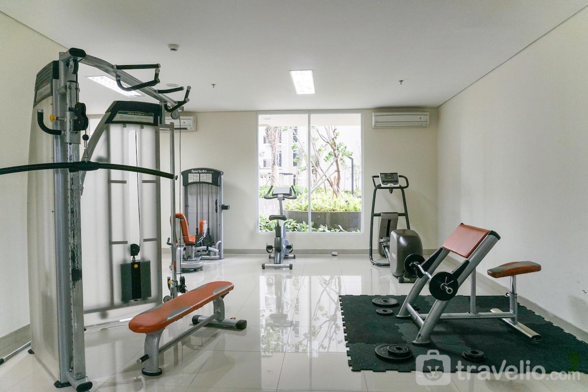 A fitness center is presented with modern workout equipment, including weight machines and a bench for strength training. Large windows allow natural light to flood the space, offering views of greenery outside. A clean, well-maintained floor accommodates various exercise activities.