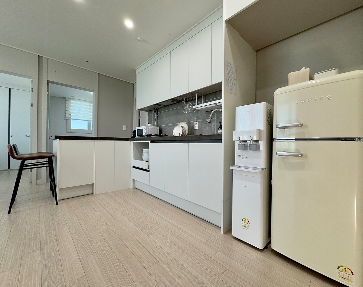 The kitchen features modern white cabinetry and sleek countertops. An eye-catching vintage-style refrigerator complements a water dispenser. Essential appliances such as a microwave and stove are included, along with seating available at a small dining area. Natural light enters through a nearby window.