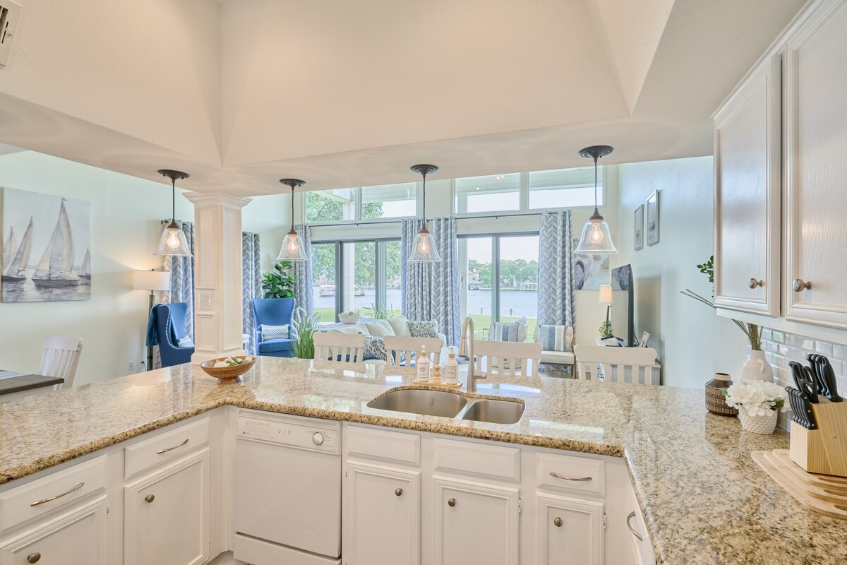 The kitchen features light-colored cabinetry and a spacious granite countertop with a double sink. Pendant lights hang above, and an open view reveals a comfortable living area with large windows overlooking the lake. Natural light enhances the bright, airy atmosphere.