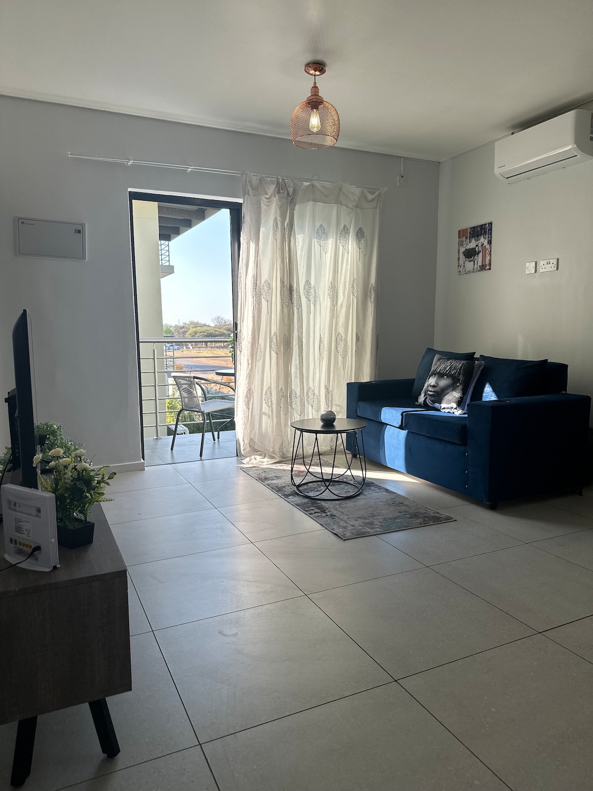 A well-lit living area features a dark blue sofa positioned next to a small coffee table. Sheer curtains drape over a spacious window, allowing natural light to fill the space. A decorative plant sits on the table, adding a touch of greenery.