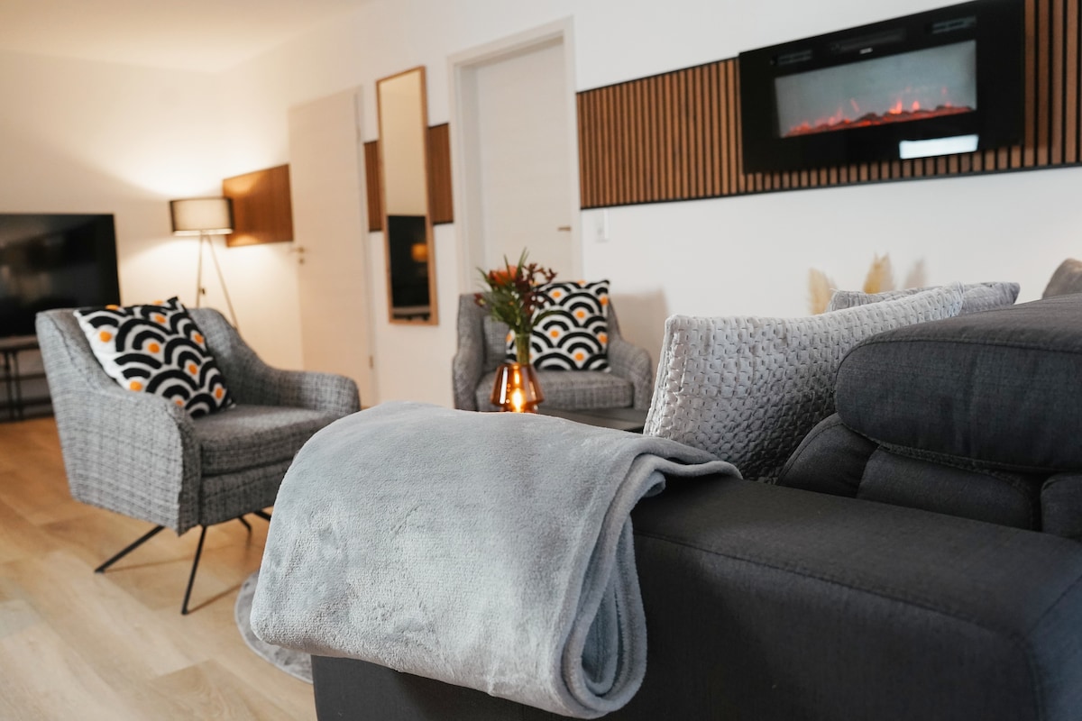 The living area features two stylish armchairs with patterned cushions facing a modern wall-mounted electric fireplace. A soft blanket is draped over the couch, enhancing the cozy atmosphere. A flat-screen television is visible in the background, contributing to the welcoming space.
