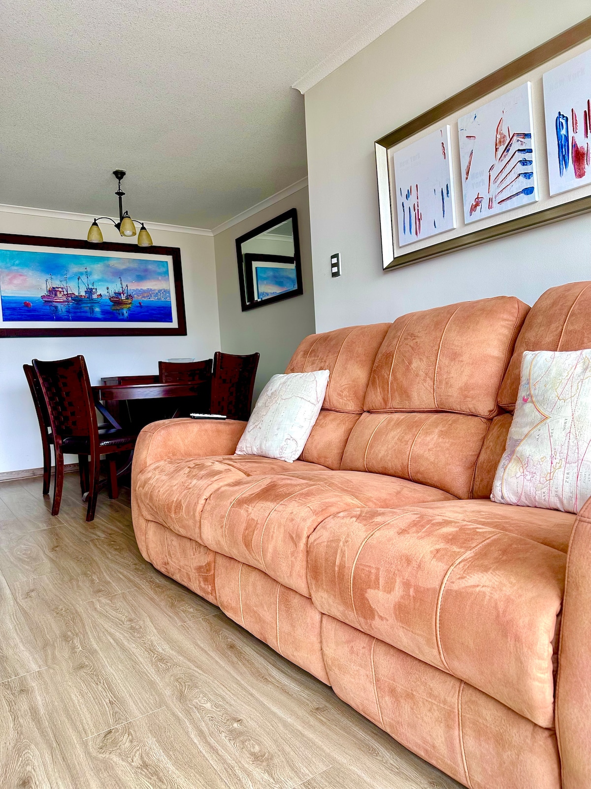 A plush, light-brown sofa is positioned in the living area, featuring decorative pillows. In the background, a dining table with dark wooden chairs is visible, accompanied by framed artwork on the wall that depicts a coastal scene.