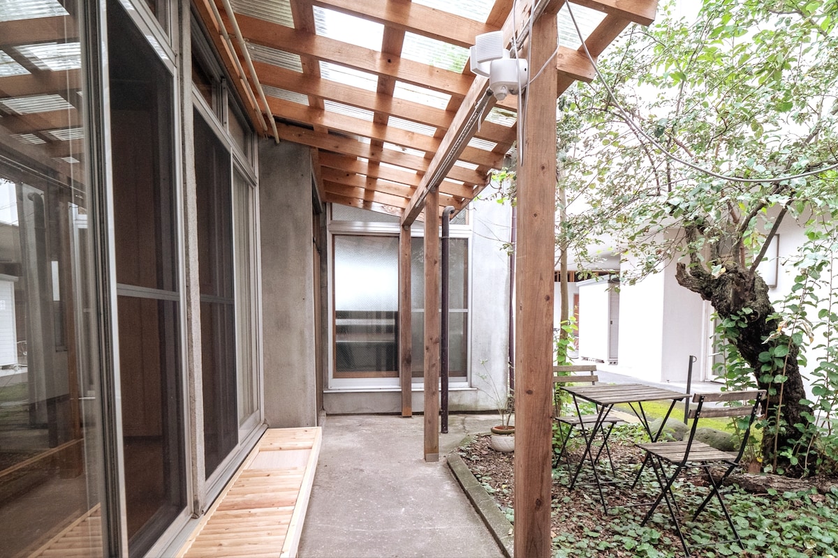 An outdoor space is featured, showcasing a wooden pergola that provides a shaded area. A small table with folding chairs is positioned on the concrete floor, surrounded by greenery. Large glass doors lead to the interior, enhancing the sense of openness.