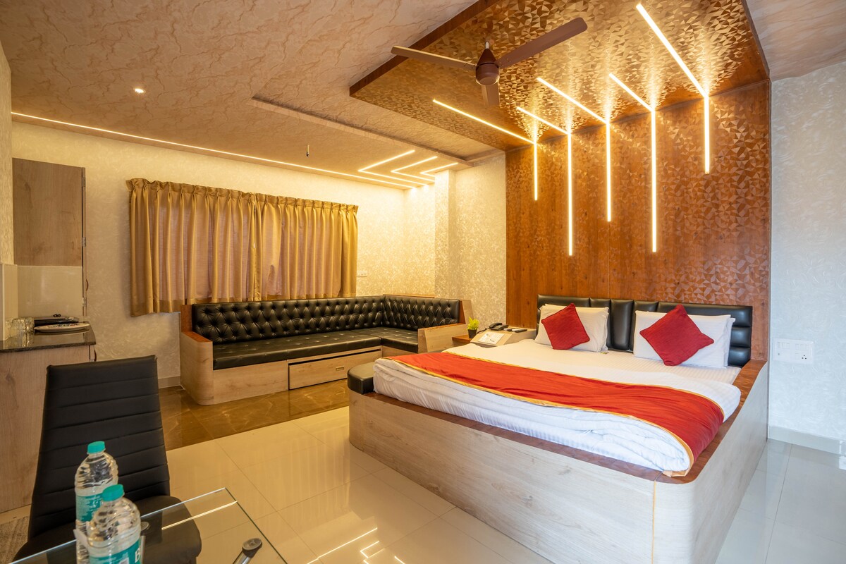 A spacious room features a king-sized bed adorned with a vibrant red throw. A sleek seating area with a black leather sofa encourages relaxation. Warm lighting highlights the textured wooden ceiling, while large curtains frame the ample windows, creating a well-lit and inviting environment.