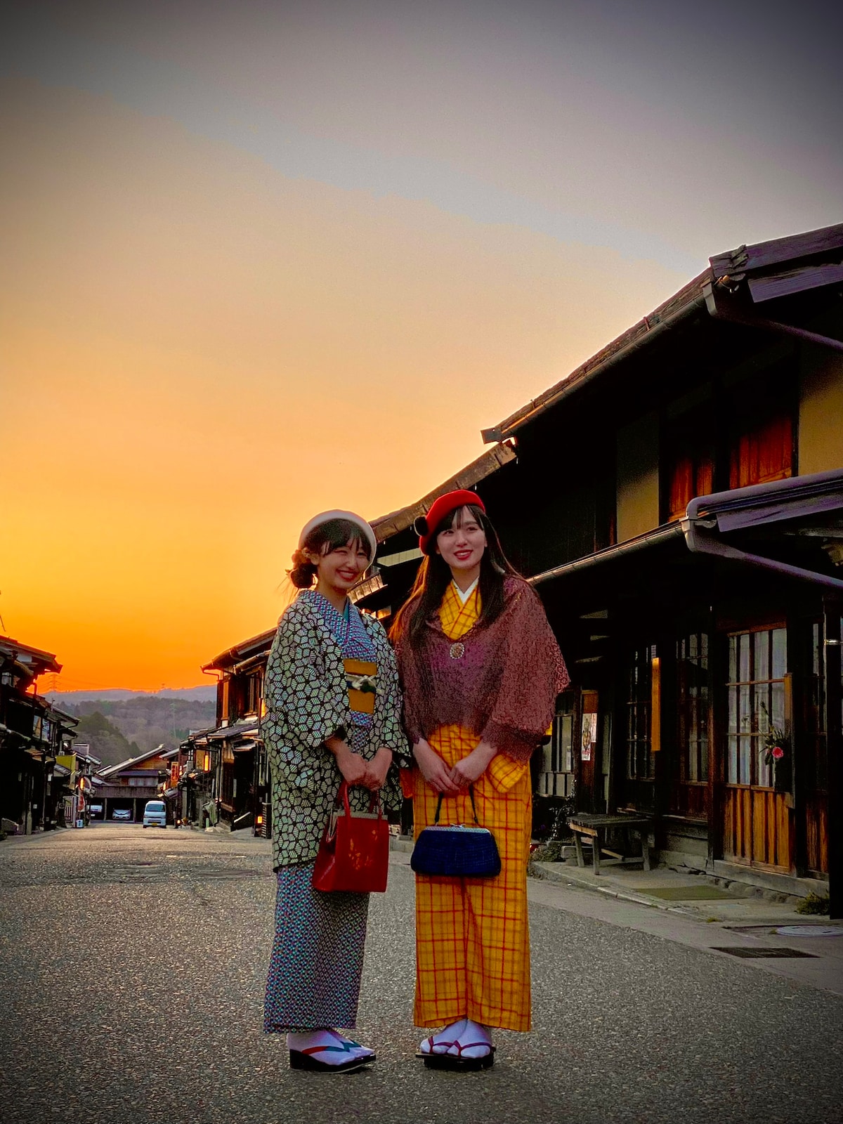 Two individuals are seen standing on a quiet street lined with traditional wooden buildings during sunset. The vibrant colors of the sky create a warm backdrop, while the architecture highlights the nostalgic charm of the surrounding area.