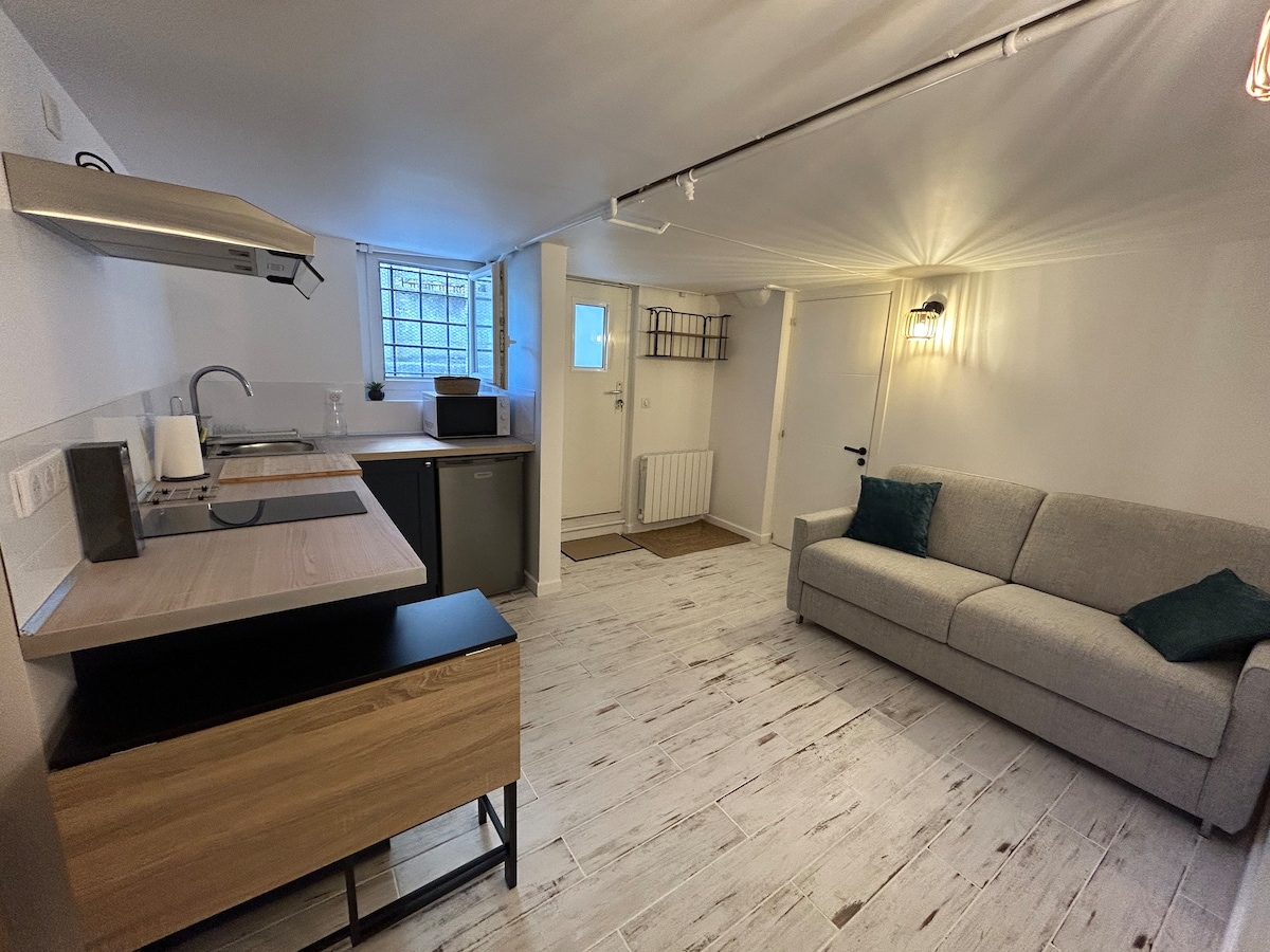 A modern studio kitchen is featured with a sleek countertop and basic appliances, including a sink and refrigerator. A well-lit living area contains a comfortable gray sofa. A separate entrance is visible with a window that allows natural light, enhancing the space's airy feel.
