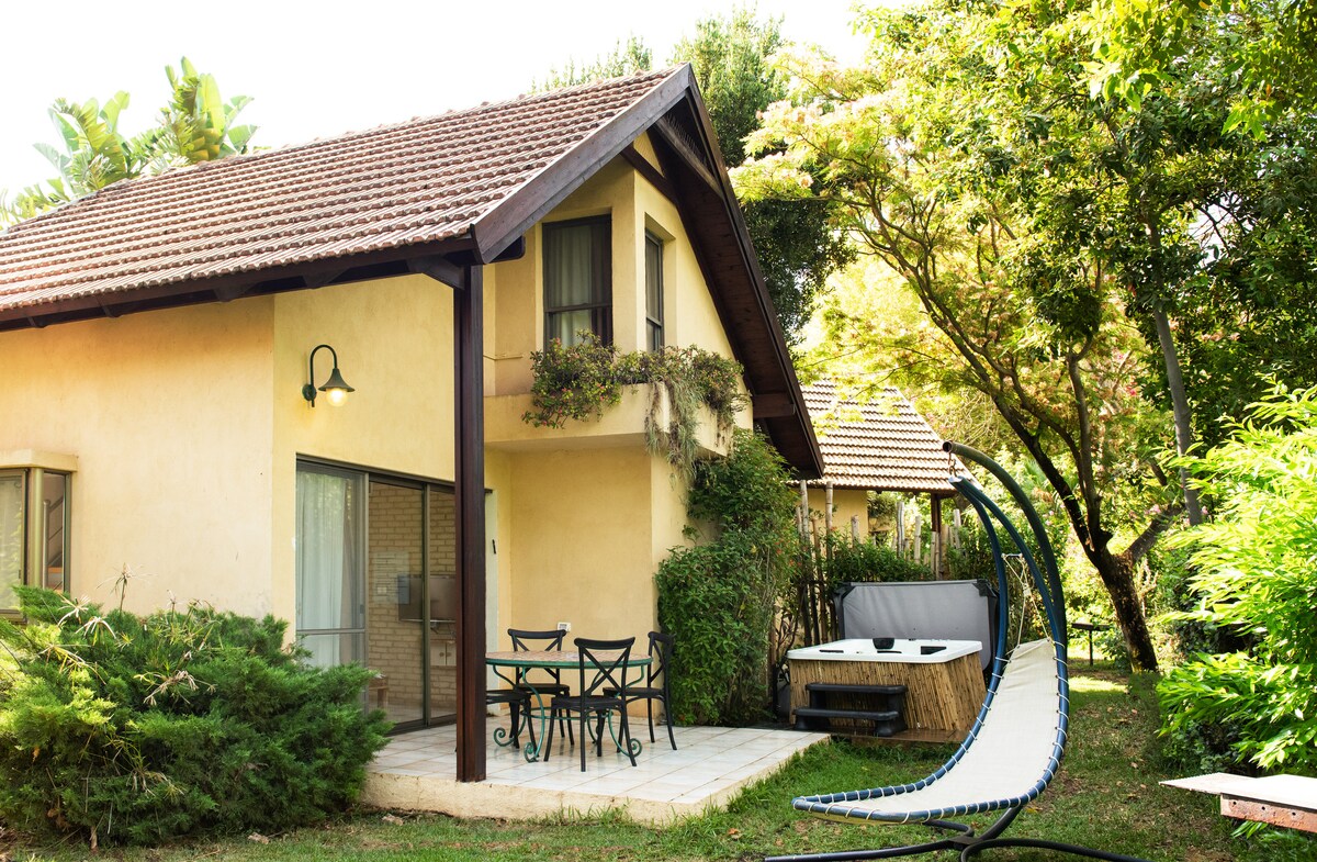 A two-story vacation home is set in a lush garden, featuring a patio with a table and chairs. A hammock is positioned nearby, while a spacious jacuzzi is visible on one side, complemented by various greenery that enhances the peaceful surroundings.