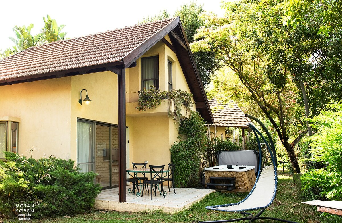 A charming exterior of the house is framed by lush greenery. A patio area features a small table and chairs, inviting relaxation. A hammock is positioned nearby, enhancing the serene outdoor space, while a jacuzzi is nestled discreetly under the shade of trees.