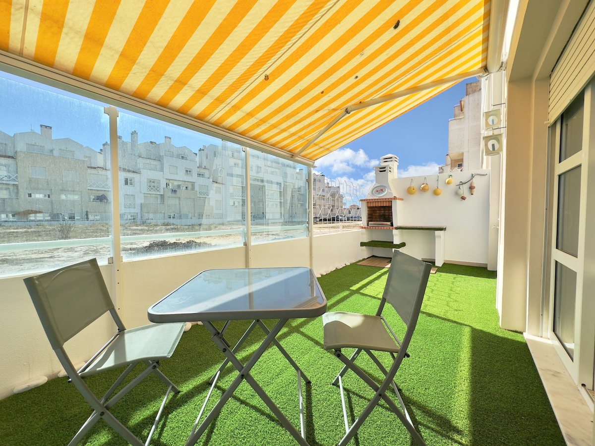 A terrace is presented with a striped yellow and white awning, offering shade over a glass-top table and two green folding chairs. The area features artificial turf, creating a green space, with a barbecue and outdoor kitchenette visible in the background.