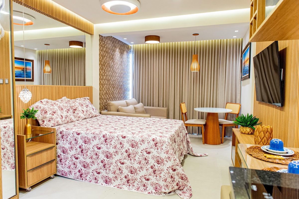 A cozy studio space features a spacious bed adorned with a floral patterned coverlet. Mirrors reflect soft lighting, and a comfortable seating area with a small dining table is visible. Neutral tones create a calm ambiance, highlighted by decorative plants and modern furnishings.
