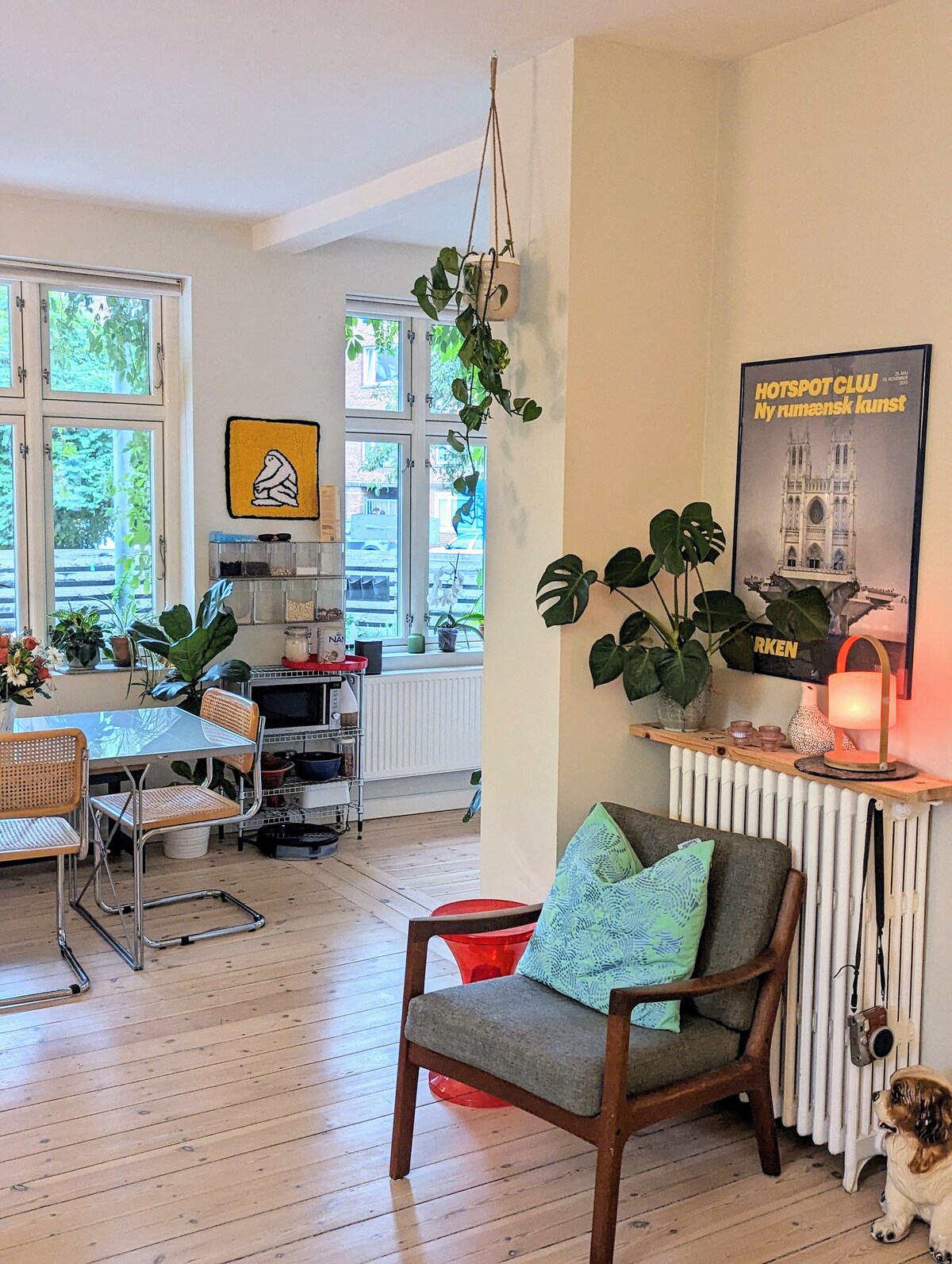 A light-filled living area showcases large windows surrounded by greenery. A dining table with metal chairs is present, alongside a cozy chair and decorative plants. An art poster is mounted on the wall, and a small lamp casts a gentle glow on a radiator.