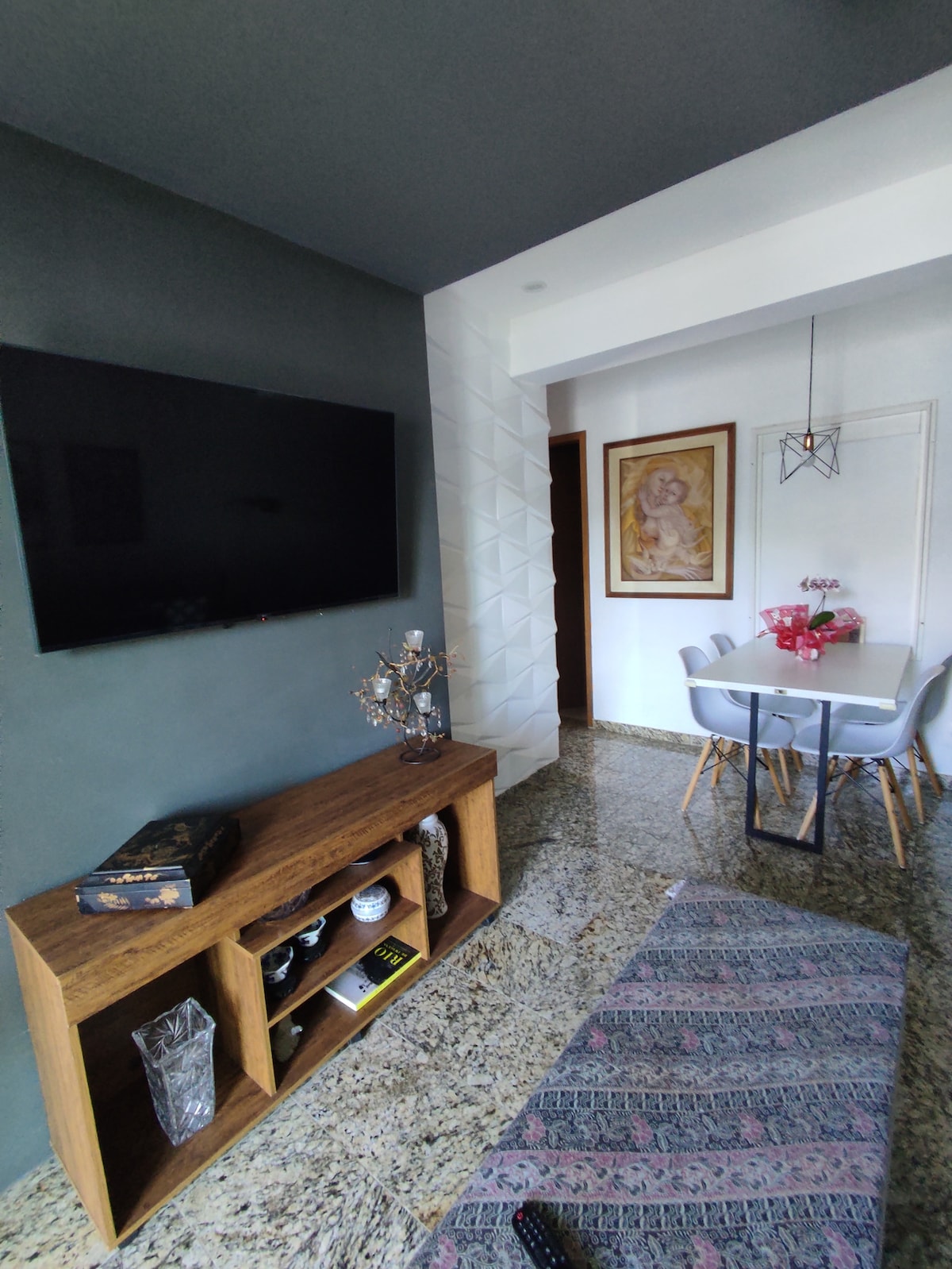 A modern living area features a large flat-screen television mounted on a textured wall. A wooden sideboard displays decorative items. Nearby, a dining table with four chairs is set with a vase of pink flowers, creating a welcoming and organized environment.