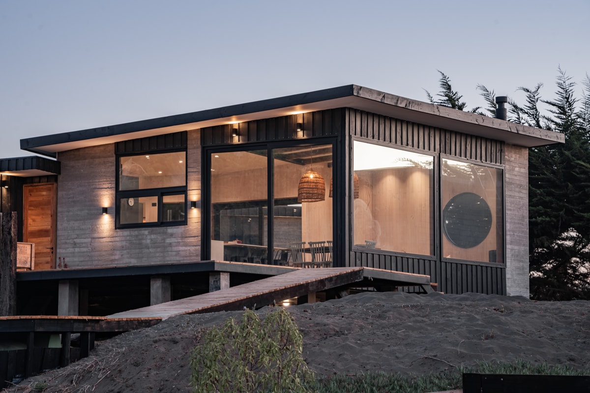 The exterior of a modern house is showcased, featuring large glass windows and a wooden facade. A sloped pathway leads to the entrance, illuminated by warm outdoor lighting. Surrounding greenery contributes to a tranquil environment, with the beach visible beyond the structure.