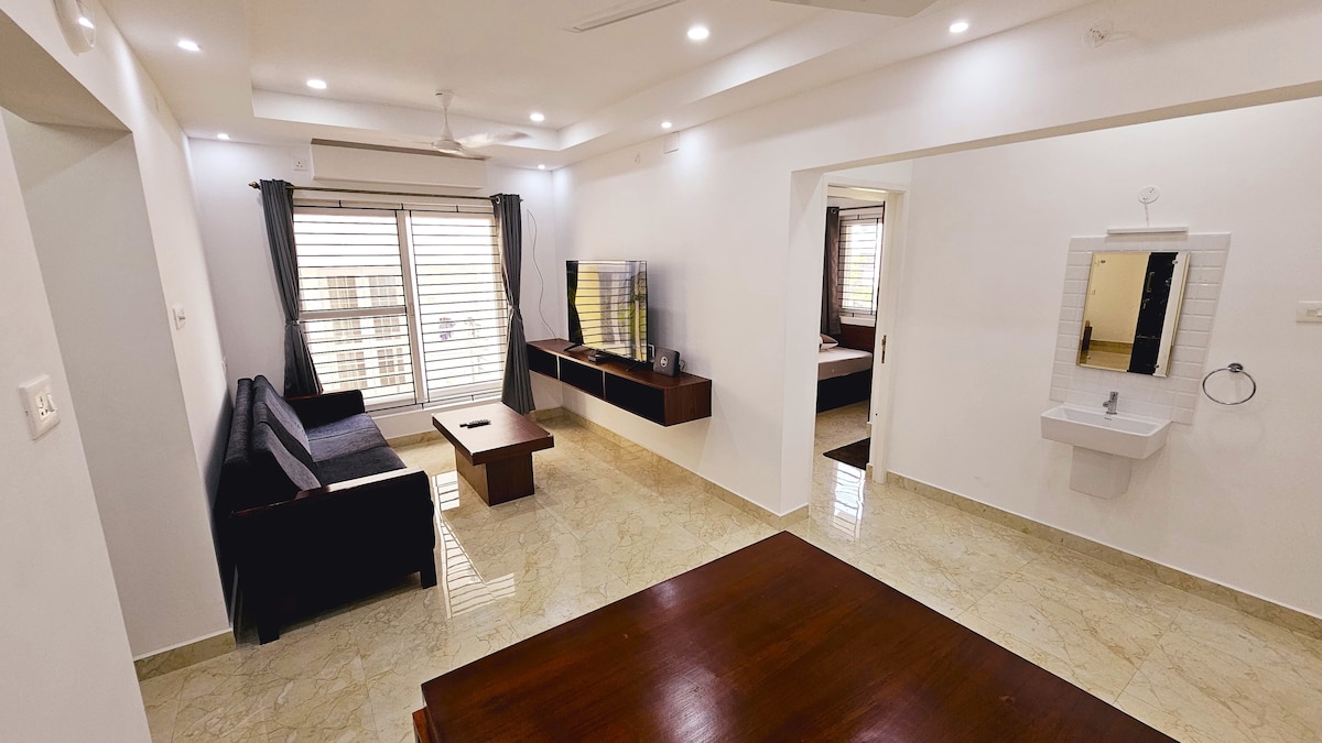 A spacious living area is presented, featuring modern furnishings including a dark sofa and a wooden coffee table. Natural light enters through large windows with simple drapes. A television is mounted on the wall, complemented by light-colored marble flooring and minimalistic decor.