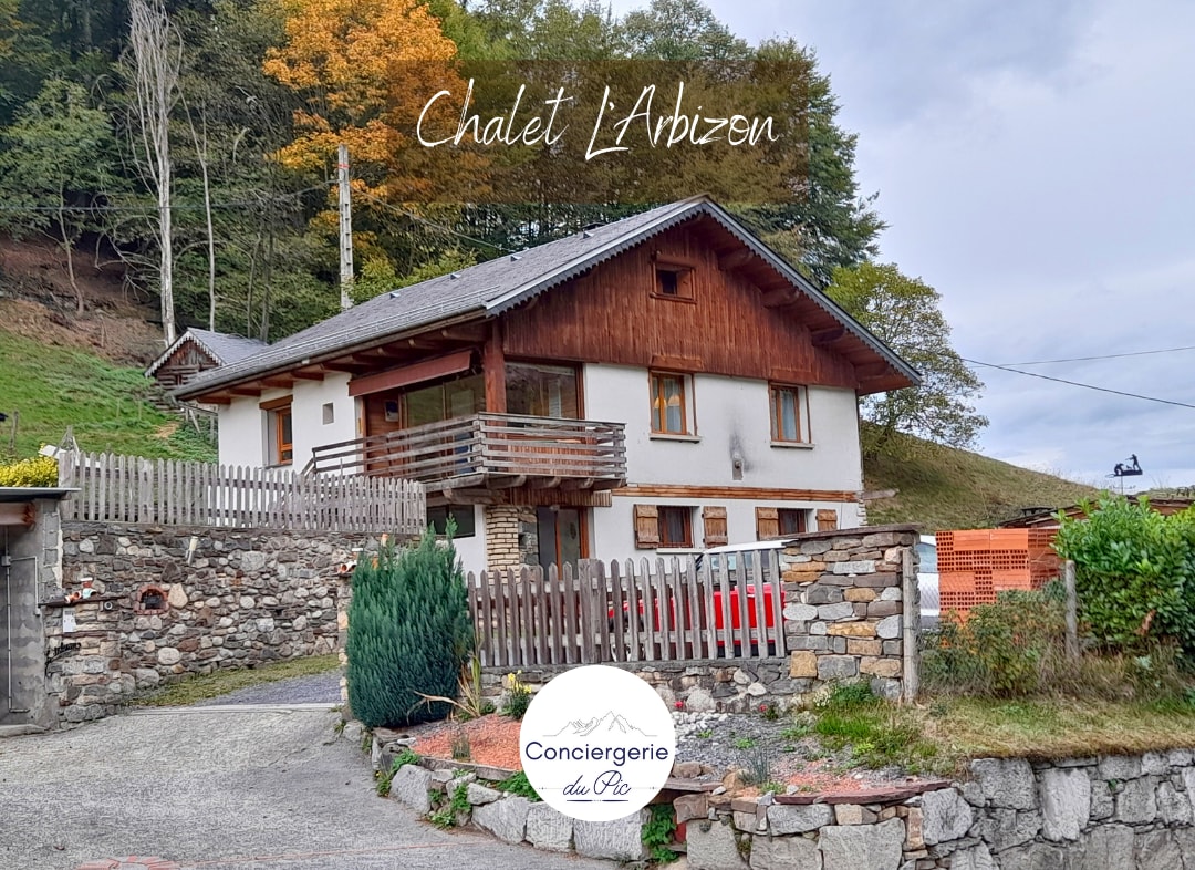 The exterior of the chalet is presented, showcasing a two-story structure surrounded by greenery. A wooden fence encloses the entrance, and colorful autumn foliage is visible in the background. The entrance features a covered porch, leading into a welcoming atmosphere.