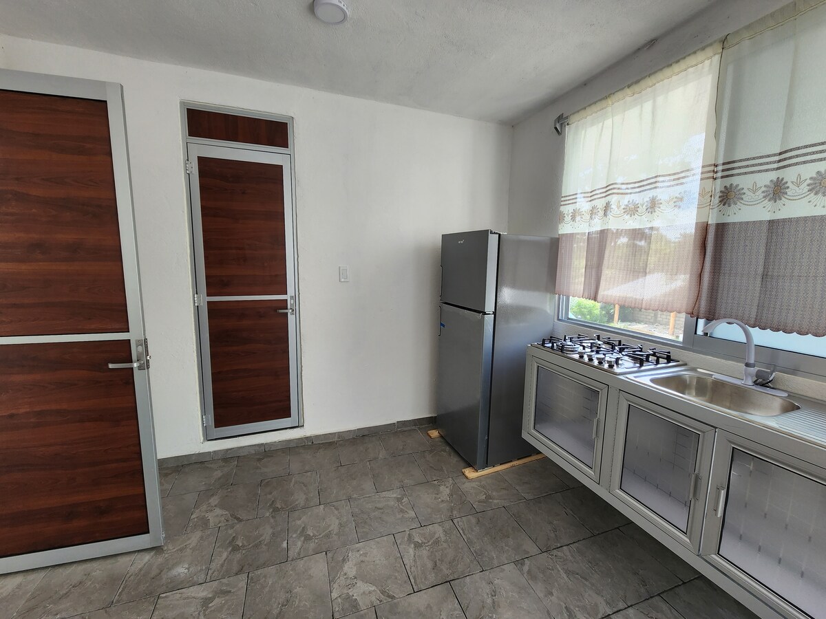 A bright kitchen area features a stainless steel refrigerator and a gas stove. Counter space is available beside a window covered with sheer curtains, allowing natural light to fill the room. Two wooden doors provide access to other areas of the accommodation.