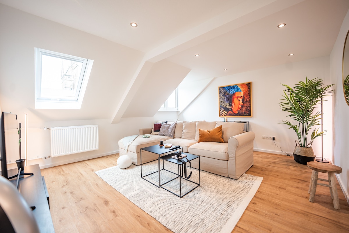 A bright and airy living room features a modern light-colored sectional sofa positioned around a chic coffee table. Notable elements include a vibrant piece of wall art, a potted plant, and a soft area rug, while angled rooftop windows allow natural light to enhance the space.