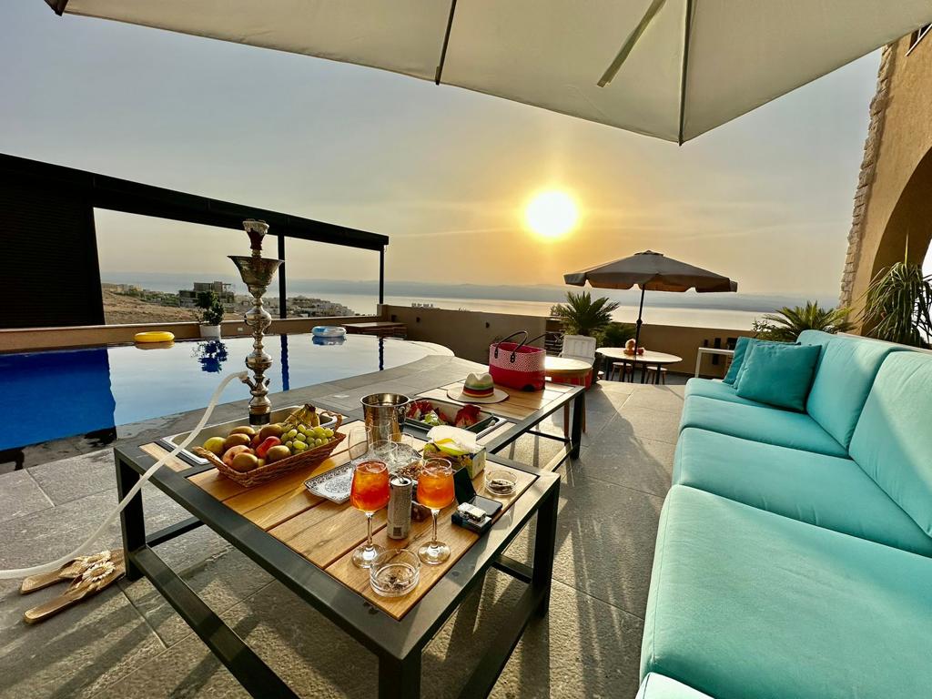 A spacious outdoor seating area is adorned with a comfortable turquoise couch and a wooden table holding a variety of fruit and beverages. A sun umbrella provides shade, while the sun sets over the horizon, casting a warm glow on the infinity pool beyond.