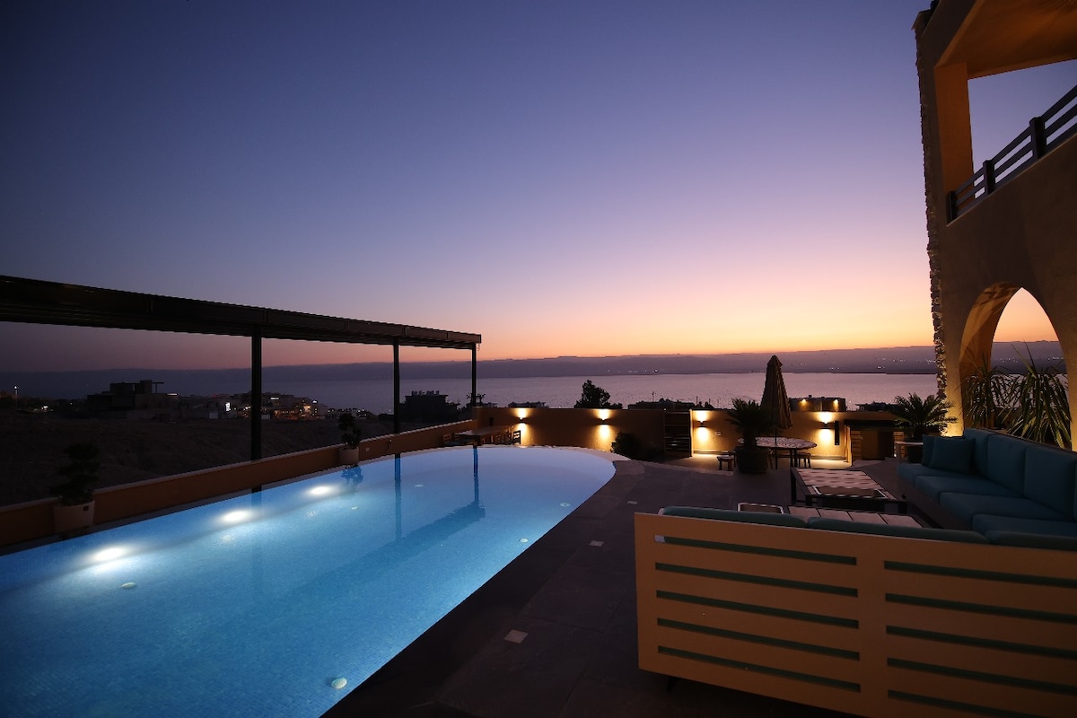A serene outdoor area features a spacious infinity pool, reflecting the colors of the twilight sky. Comfortable seating is arranged nearby, and the horizon showcases the tranquil waters of the Dead Sea. Subtle lighting enhances the overall ambiance.