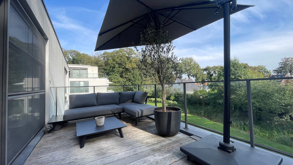 A large terrace features modern outdoor furniture, including a sectional sofa and a coffee table. A large umbrella provides shade. Lush greenery from the landscaped garden is visible, enhancing the outdoor space's relaxed atmosphere.