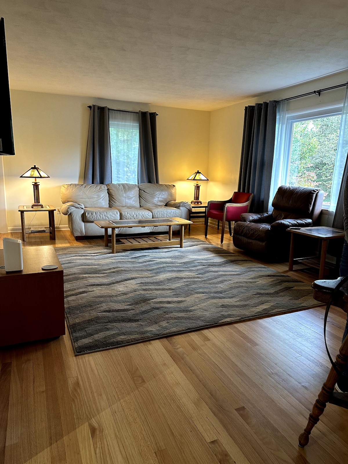 The living area features a light-colored sofa paired with a spacious coffee table situated on a patterned area rug. Two accent chairs, one red and one brown, complement the seating arrangement. Natural light is provided by a nearby window, framed by dark curtains.
