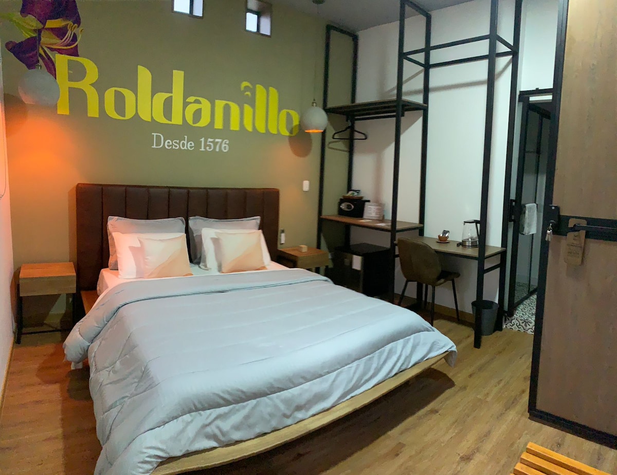 A cozy room is highlighted by a spacious bed with soft linens and pillows. A wooden headboard complements the modern decor. A small workspace with a chair is visible, along with shelf space and a hanging rack. Natural light is provided by multiple windows.