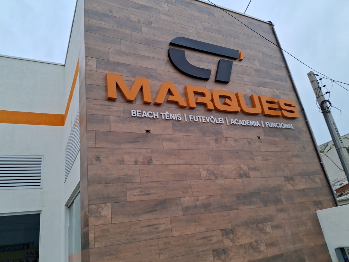 The exterior of a modern building showcases the prominent signage of 'MARQUES' in bold, orange lettering. The sign highlights offerings such as Beach Tênis, Futevôle, Academia, and Functional training. A sleek wood-patterned façade complements the contemporary design.