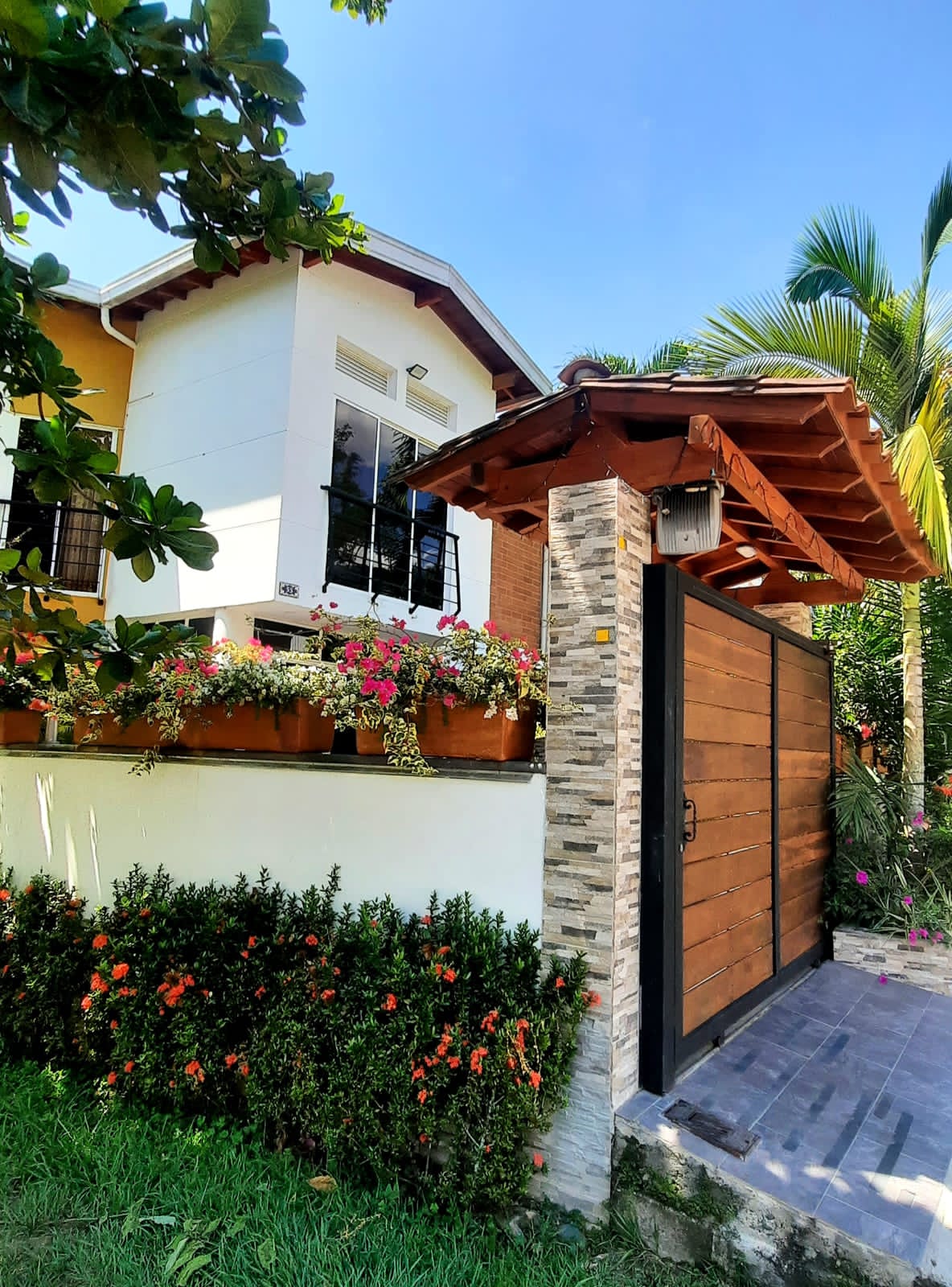 The entrance features a wooden gate adorned with stone accents, surrounded by colorful flowering plants and lush green grass. The house is visible in the background, showcasing a charming architectural style with large windows and a welcoming porch.