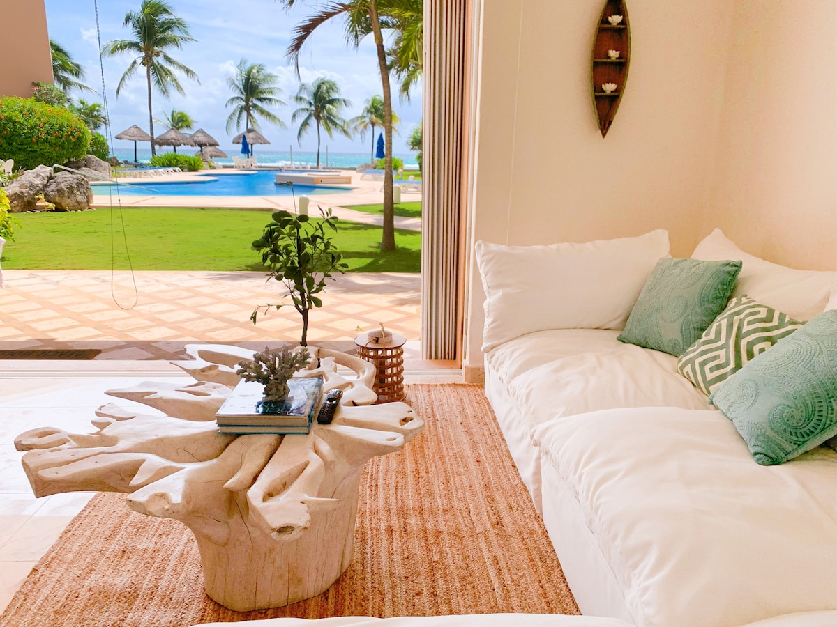 A cozy seating area features a light-colored sofa adorned with decorative pillows. A unique coffee table carved from natural material rests on a textured area rug. Tropical plants and views of the pool and ocean are visible through expansive glass doors.