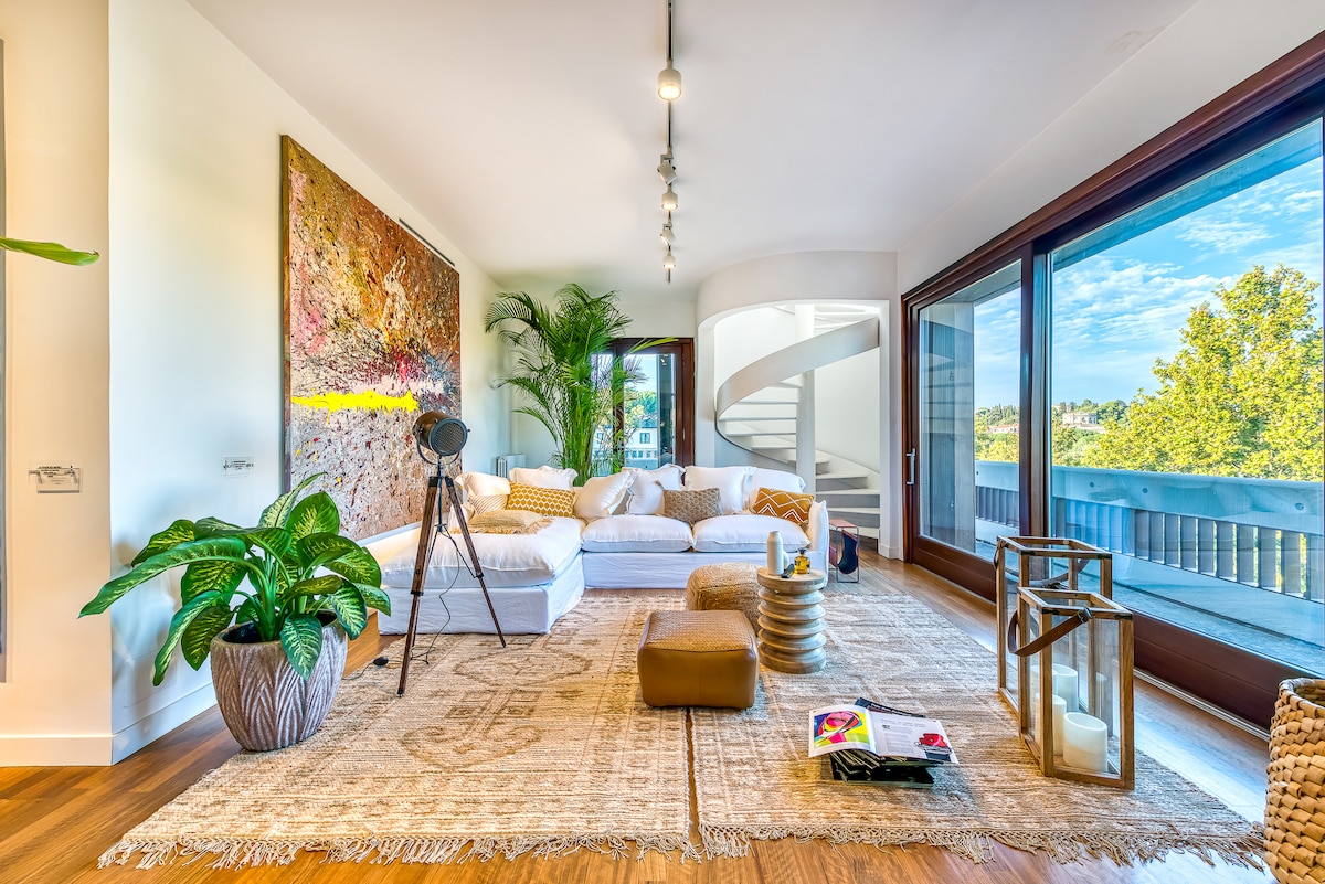 A spacious living area features a comfortable white sofa, accentuated by a colorful abstract artwork on the wall. Natural light floods the space through large glass doors, revealing lush greenery outside. A decorative plant and various stylish accessories enhance the inviting atmosphere.