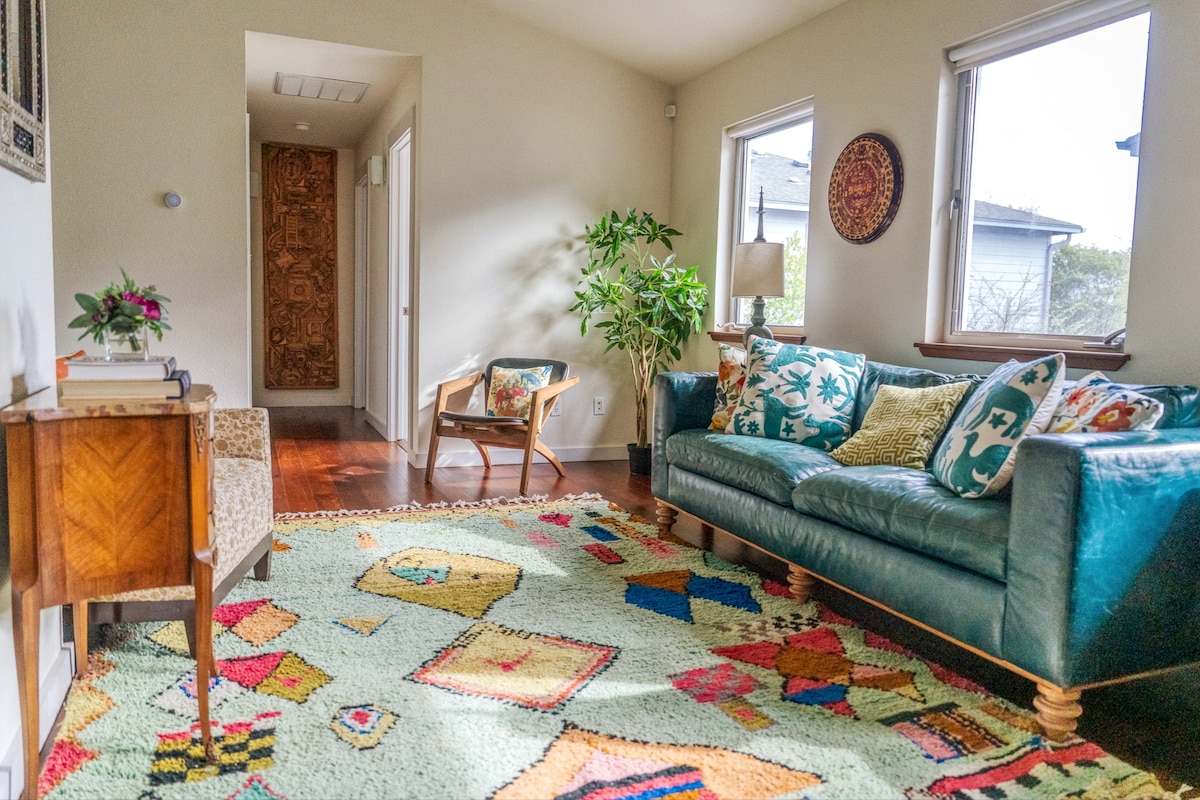 A sunlit living area is highlighted by a vibrant, patterned rug beneath a spacious green sofa adorned with decorative pillows. A small wooden table and an armchair complete the space, while natural light filters through two windows, enhancing the room's inviting atmosphere.