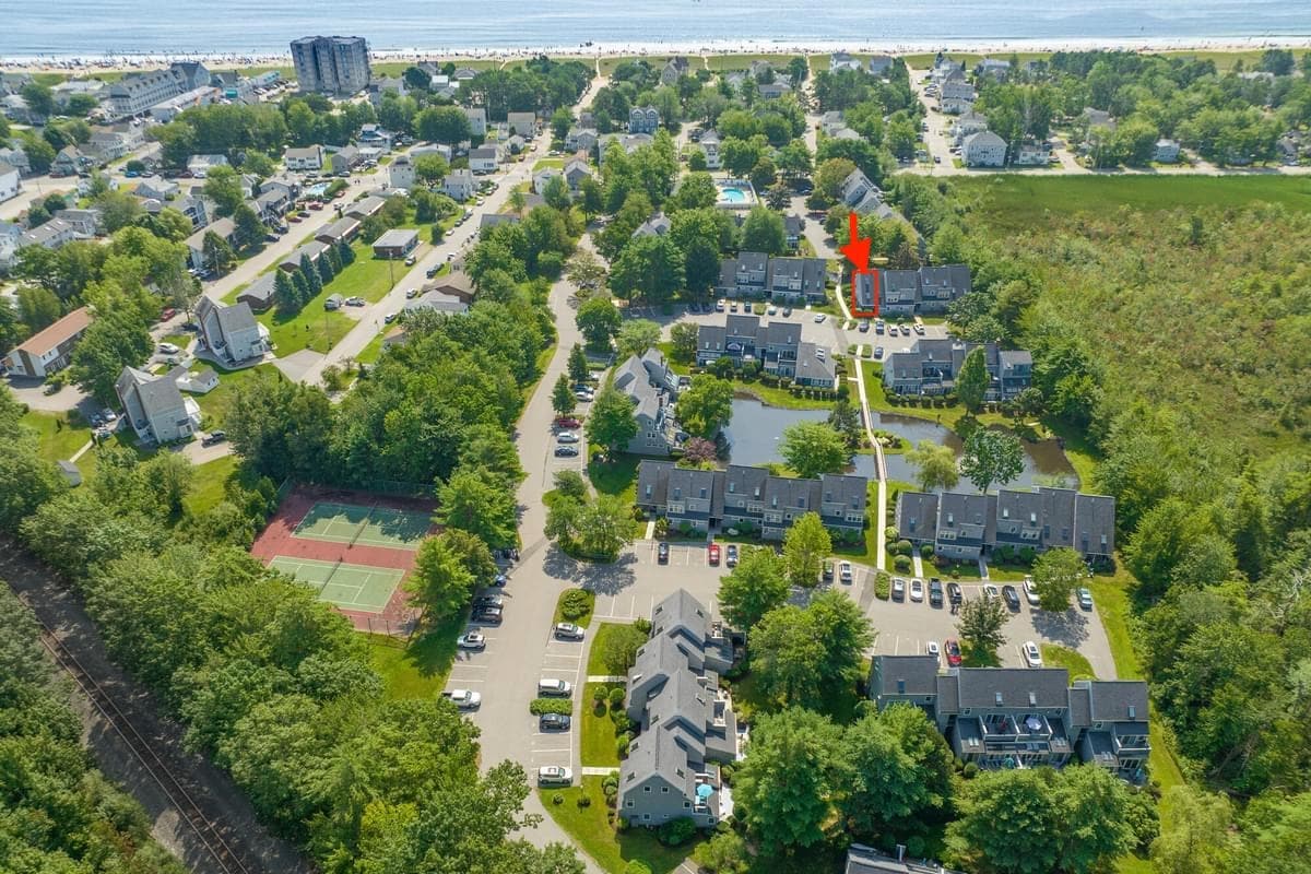 An aerial view captures the townhome complex amidst lush greenery, with tennis courts visible below. The beach lies just beyond the neighborhood, showcasing its proximity to the coast. The landscaped grounds are marked by a path leading through well-maintained gardens toward the townhomes.
