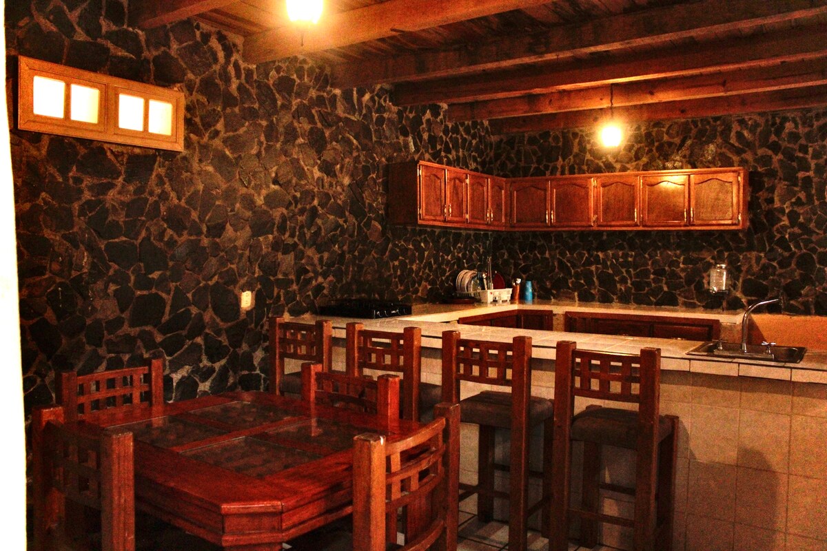 A rustic kitchen area with stone walls is featured, complemented by wooden cabinetry. A spacious dining table, surrounded by matching chairs, is positioned near the kitchen. Bar stools are arranged at the counter, enhancing the communal feel of the space.