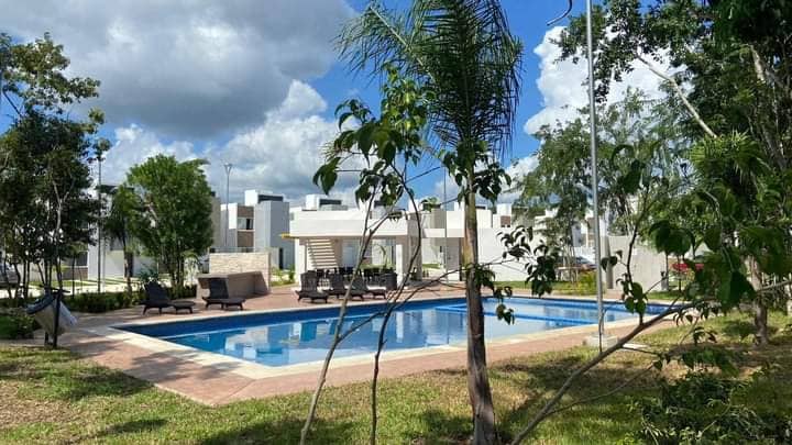 A peaceful pool area is framed by greenery, featuring a clear blue swimming pool surrounded by lounge chairs. Trees and plants add natural elements to the setting, while a well-maintained pathway leads to the entrance of the nearby accommodations.