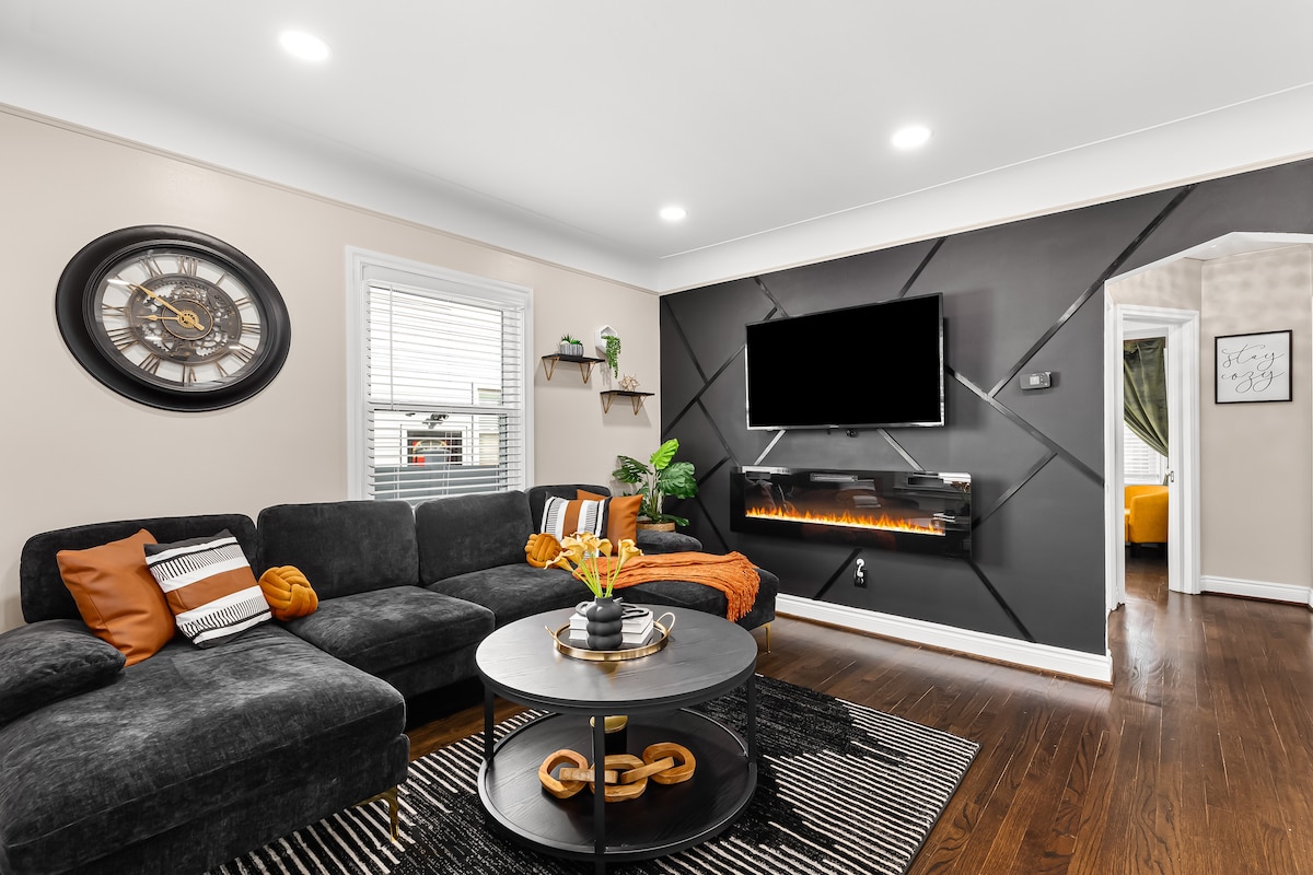 A modern living room features a dark sectional sofa adorned with orange and white pillows. A circular coffee table with decorative elements is centered in the space. A wall-mounted TV and an electric fireplace are positioned on the dark accent wall, complemented by natural light from nearby windows.