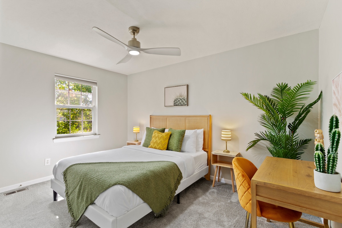 A cozy queen-sized bed with a green blanket and yellow accent pillow sits in a well-lit room. A nightstand with a lamp is positioned beside the bed, and potted plants add a refreshing touch of greenery to the space