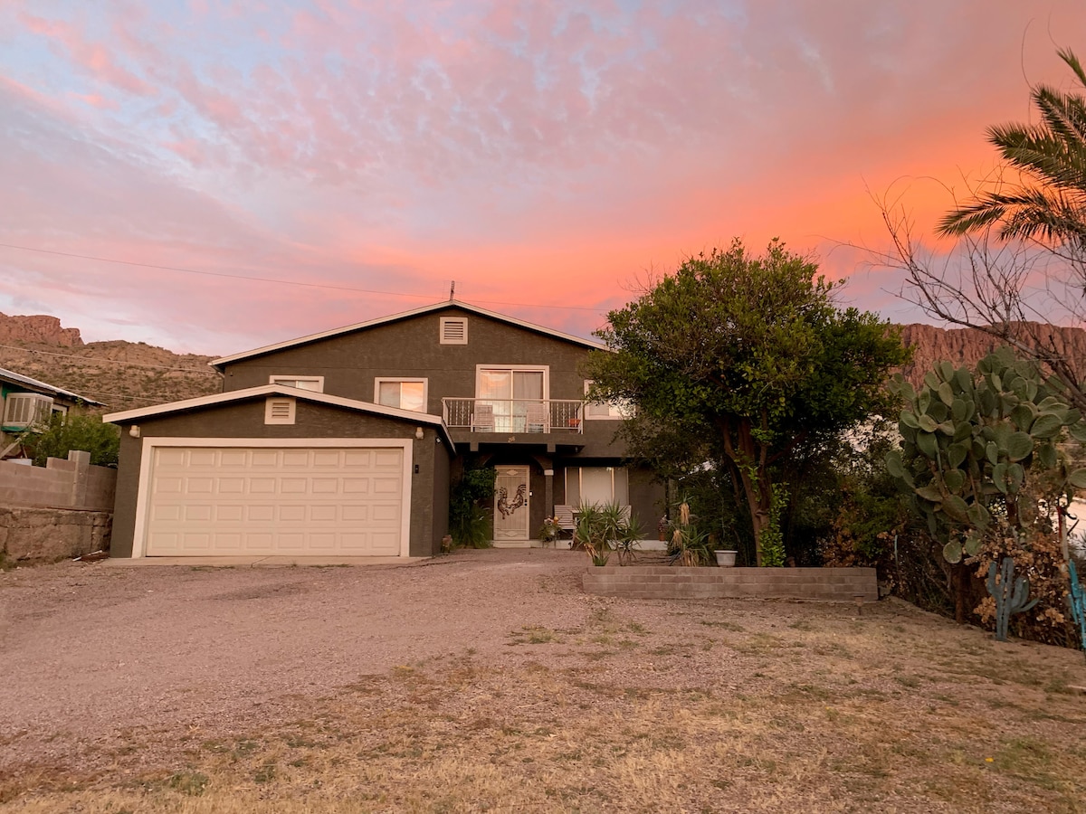 The home showcases a charming two-story design with a welcoming entrance, surrounded by desert landscaping. The exterior is complemented by vibrant sunset hues in the sky. A spacious gravel driveway provides ample parking, with cacti and greenery enhancing the setting.