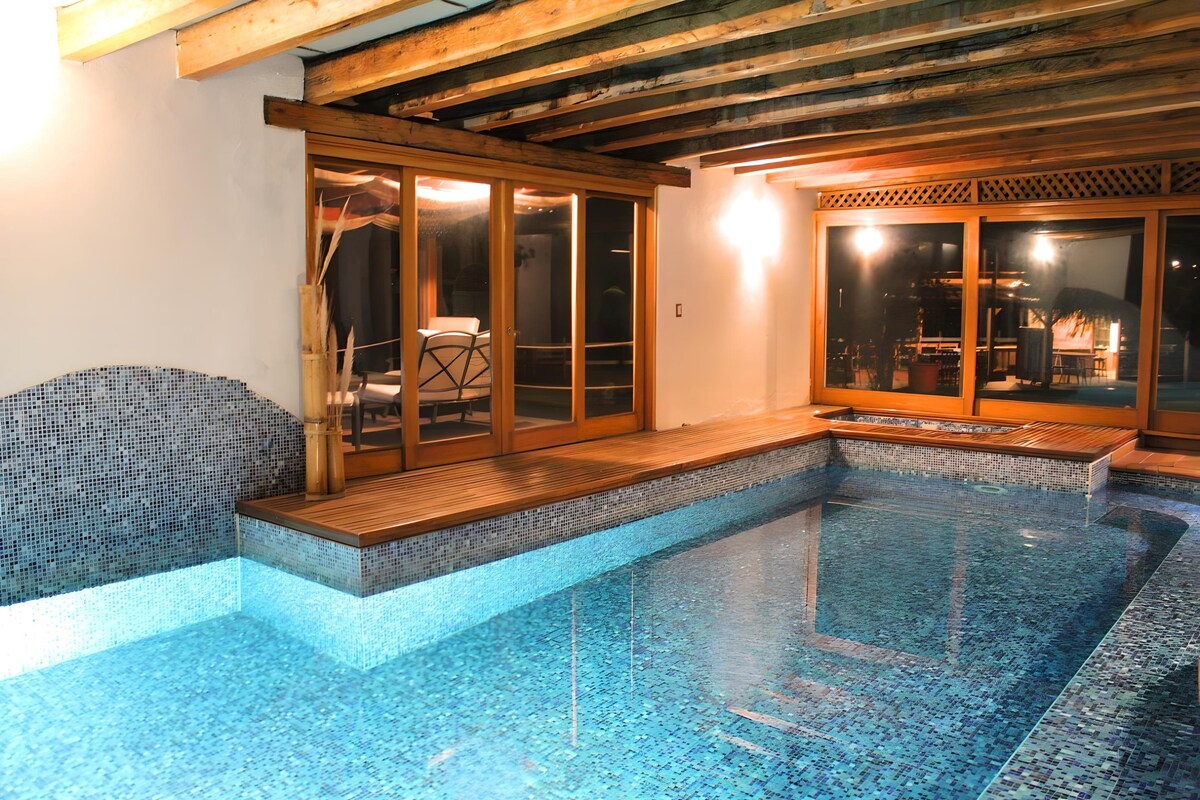 An indoor pool area is surrounded by wooden beams and large glass doors, allowing natural light to illuminate the space. The pool features mosaic tiles in shades of blue, complemented by a wooden deck. Comfortable seating is visible in the background.